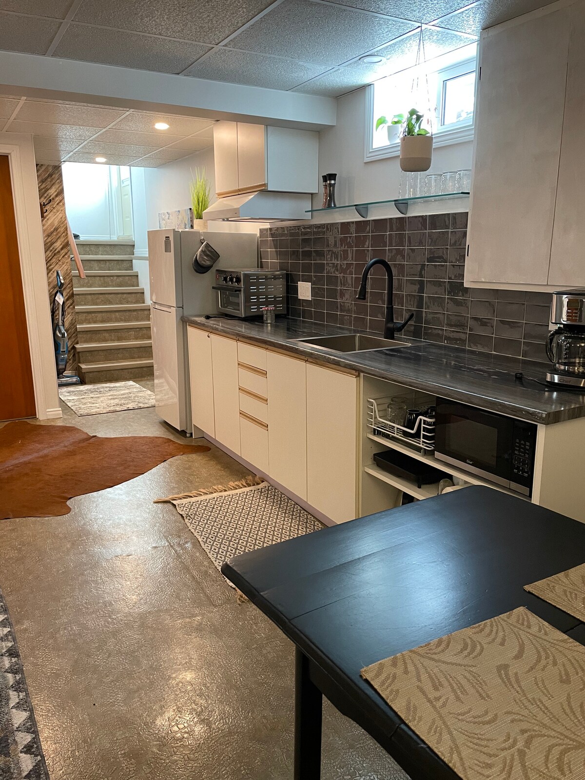 A well-equipped kitchen is visible, featuring modern appliances including a refrigerator, microwave, and stove. A black sink is centrally positioned against a stylish gray backsplash. A table is set with placemats, and textured rugs are placed atop the concrete floor, enhancing the space's functionality.