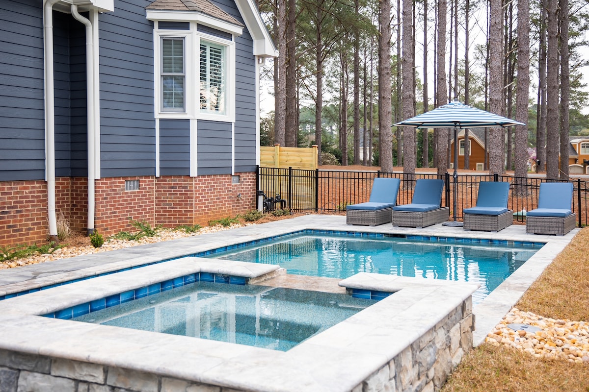 A private pool and hot tub are framed by a stone patio, featuring comfortable loungers under a blue-striped umbrella. Tall trees provide a natural backdrop, while a fully fenced yard offers privacy. The home’s blue exterior can be seen in the background.