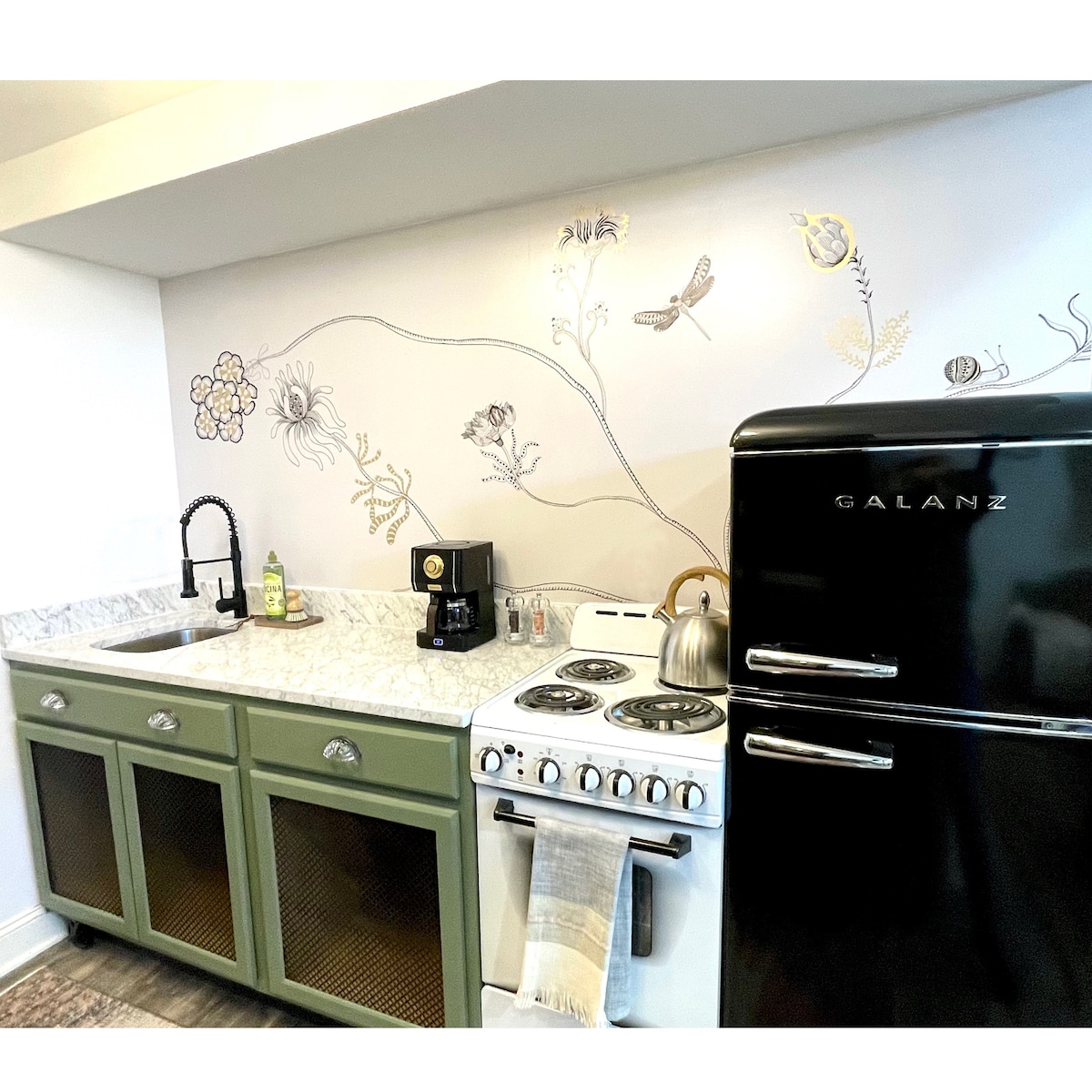 A compact kitchenette features green cabinetry with mesh doors and a stylish black refrigerator. A white countertop provides space for a coffee maker and kettle, while a vintage-style stove and a sink complete the setup. Delicate floral designs adorn the wall above.