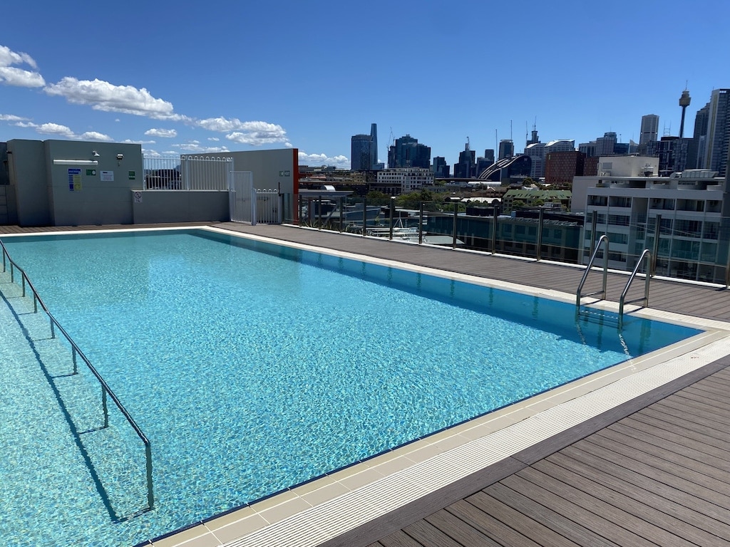 A rooftop pool is depicted, featuring clear blue water reflecting the surrounding skyline. Steps lead into the pool, while wood decking provides space for relaxation. The cityscape is visible in the background, showcasing a blend of modern buildings under a clear sky.