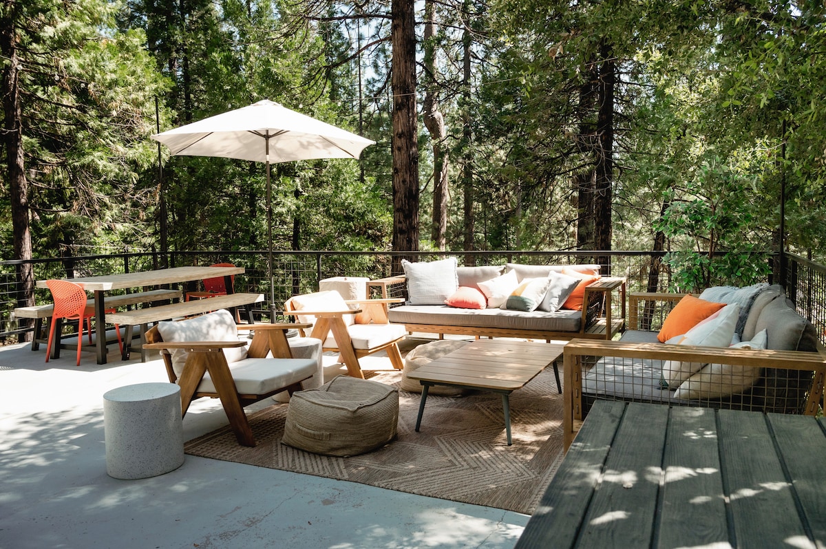 An outdoor seating area is surrounded by tall trees, featuring a large umbrella for shade. Comfortable furniture includes cushioned sofas, a low coffee table, and an additional dining table with folding chairs. A light-colored rug and soft poufs enhance the inviting space.