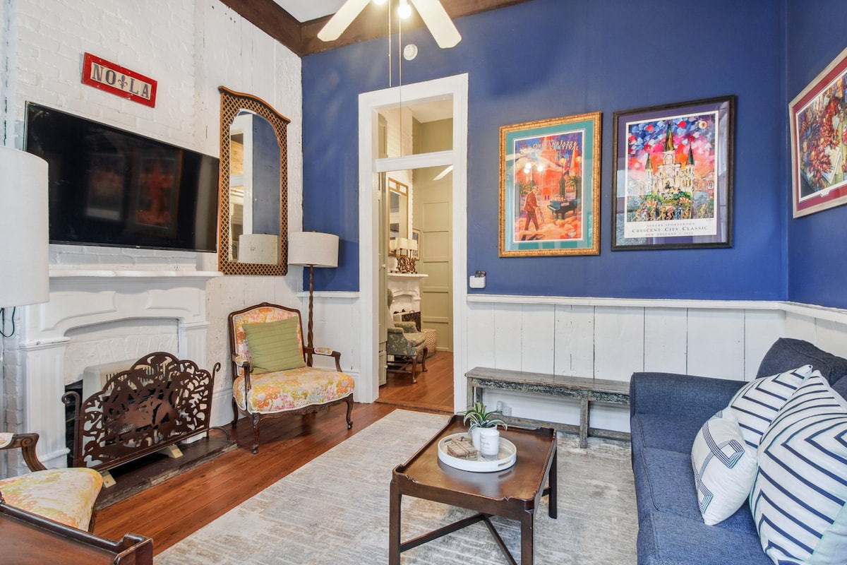 A living area features a deep blue accent wall with framed artwork. A sofa, patterned throw pillows, and a couple of chairs provide seating. A flat-screen TV is mounted above a decorative fireplace. Natural light is welcomed through an adjacent doorway.