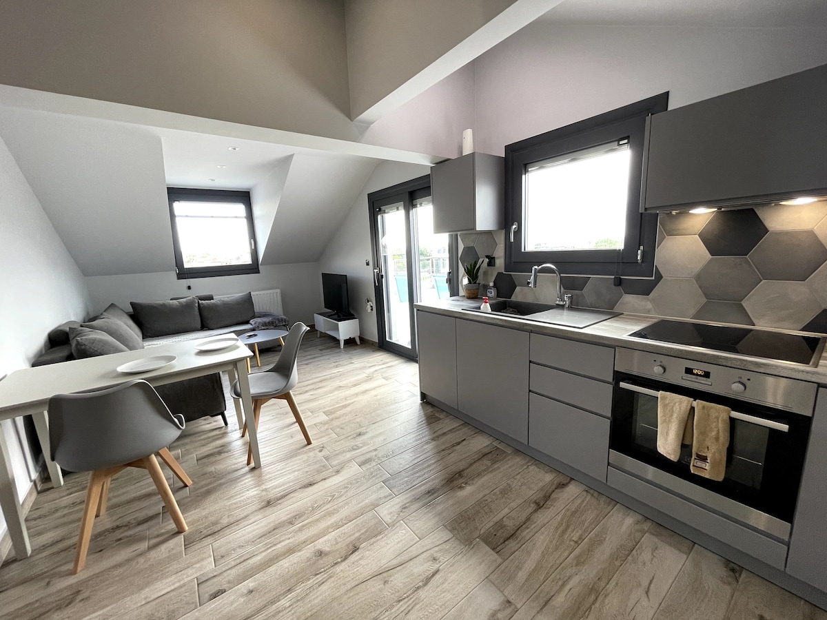 A modern kitchen area features sleek gray cabinetry and a hexagonal backsplash. A dining table with four chairs sits near the window, allowing ample natural light. A spacious living area is visible, equipped with a cozy sectional sofa and television, creating an open living space.