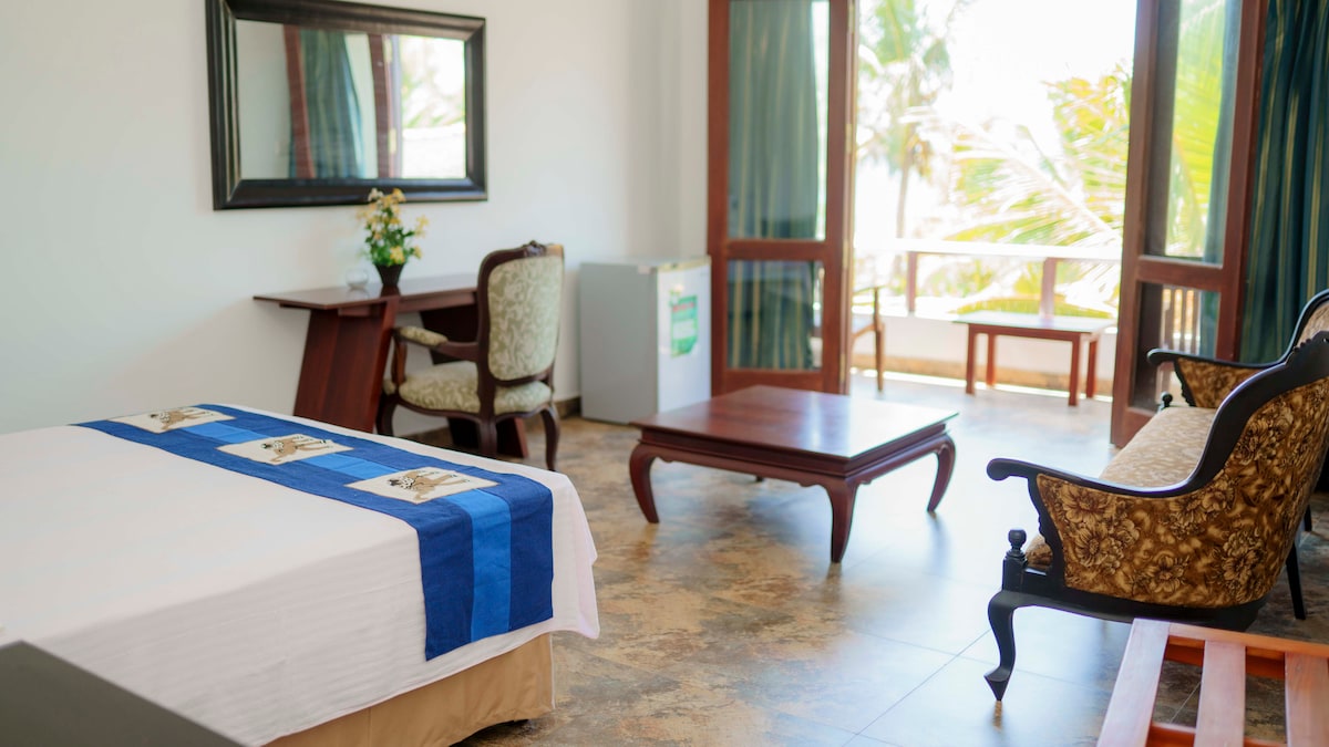 A spacious room features a large bed dressed in white linens with a blue accent. A desk and ornate armchair provide seating options. Natural light floods the area from the balcony doors, which lead to an outdoor space overlooking greenery.