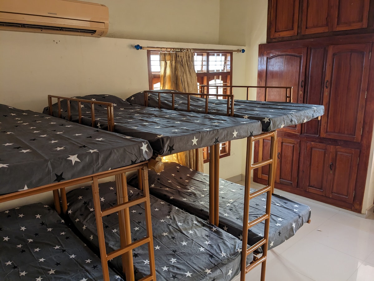 A multi-level bunk bed setup occupies the room, featuring dark fabric with star patterns. Metal frames with ladders provide access to the upper beds. Sunlight filters through the windows, illuminating the space, while wooden cabinets add a natural touch to the decor.
