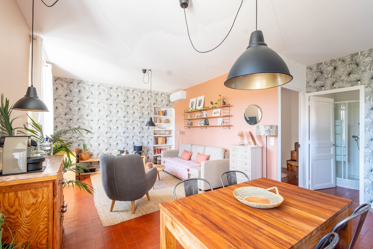A spacious living area showcases an open layout with a large window allowing natural light. A comfortable sofa sits beside a stylish armchair, while a wooden dining table is set for gatherings. Decorative elements, including plants and wall art, enhance the inviting decor.
