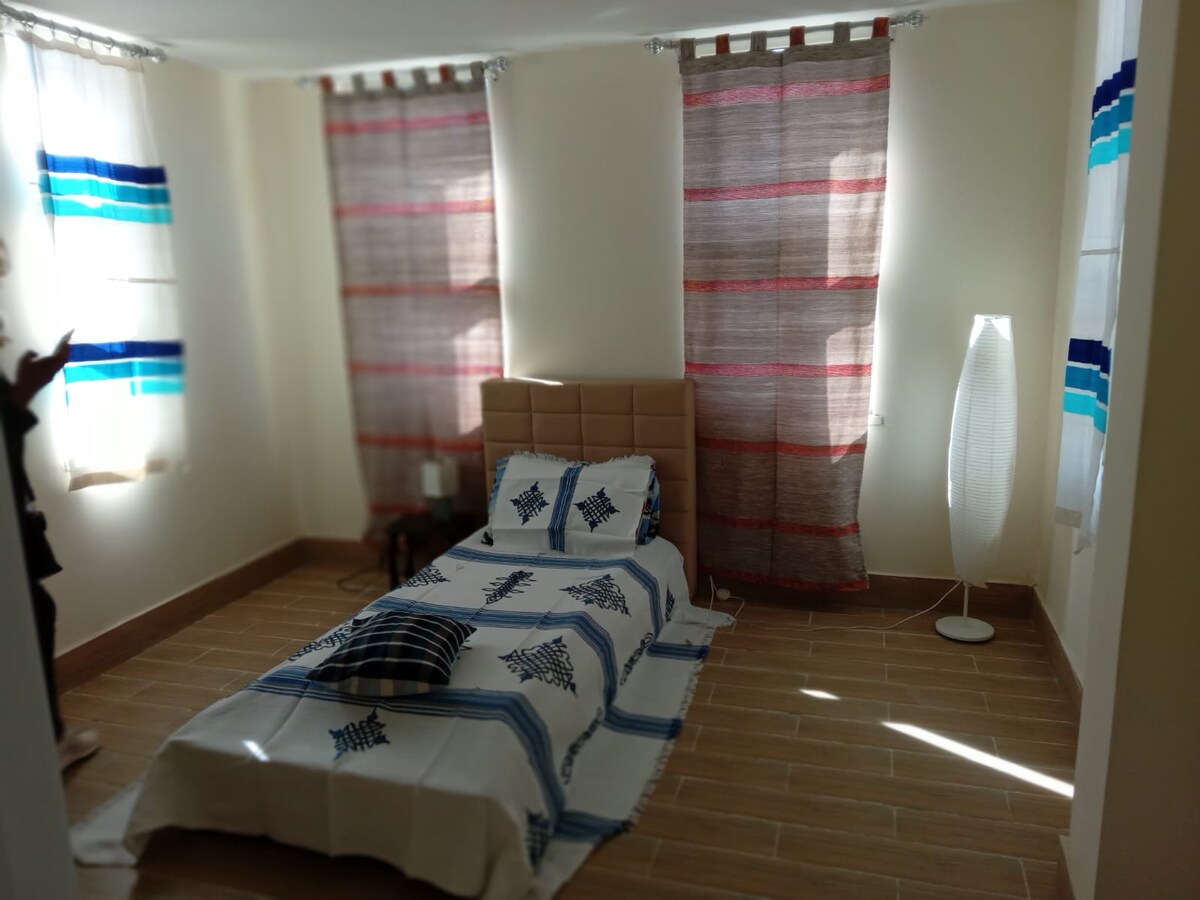 A well-lit bedroom features a spacious single bed with a patterned throw blanket. Soft sunlight filters through colorful curtains, casting gentle shadows on the tiled flooring. A slim, modern lamp stands beside the bed, enhancing the room's tranquil ambiance.