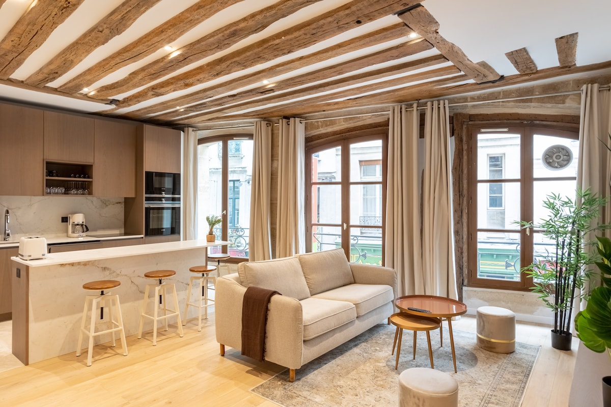 A modern living area features a spacious sofa facing large windows with natural light. Light wood beams accent the ceiling, while a kitchen bar with stools complements the open layout. Soft furnishings include a rug and side tables, enhancing the contemporary design.