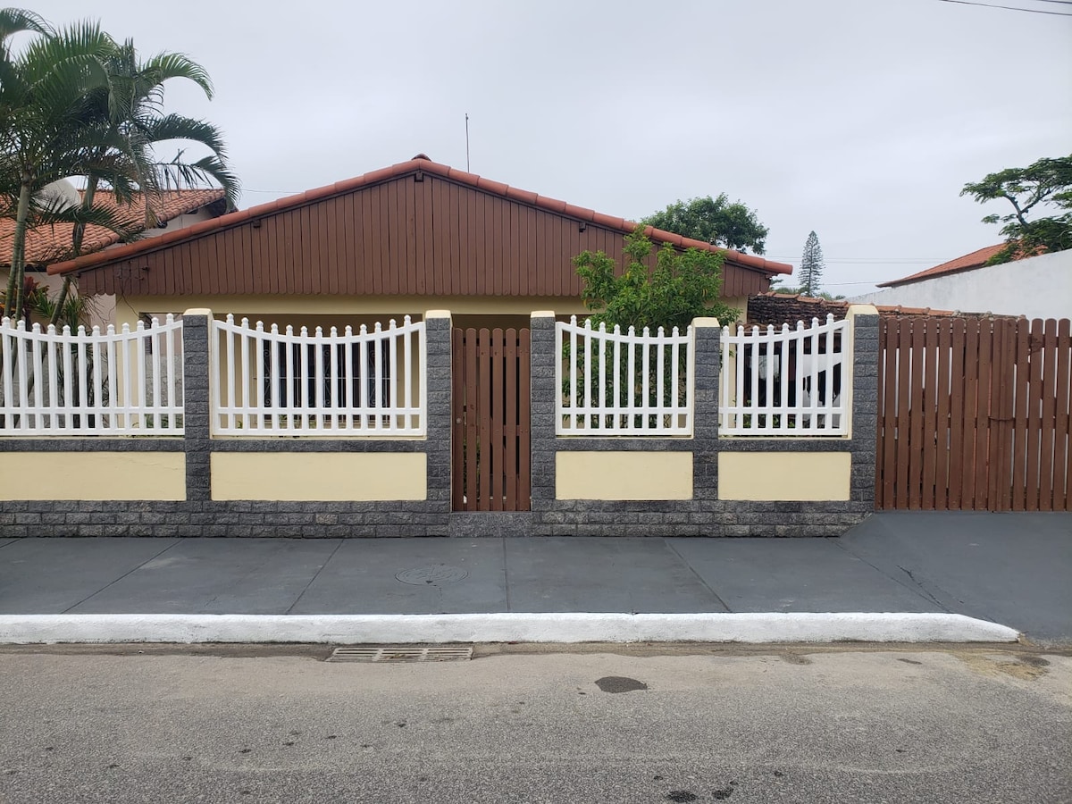 The exterior of the house features a sturdy fence with a white picket design, complemented by stone accents. A wooden gate leads to the entrance, framed by green shrubbery and palm trees. The house has a sloped roof and a welcoming façade, situated along a quiet street.