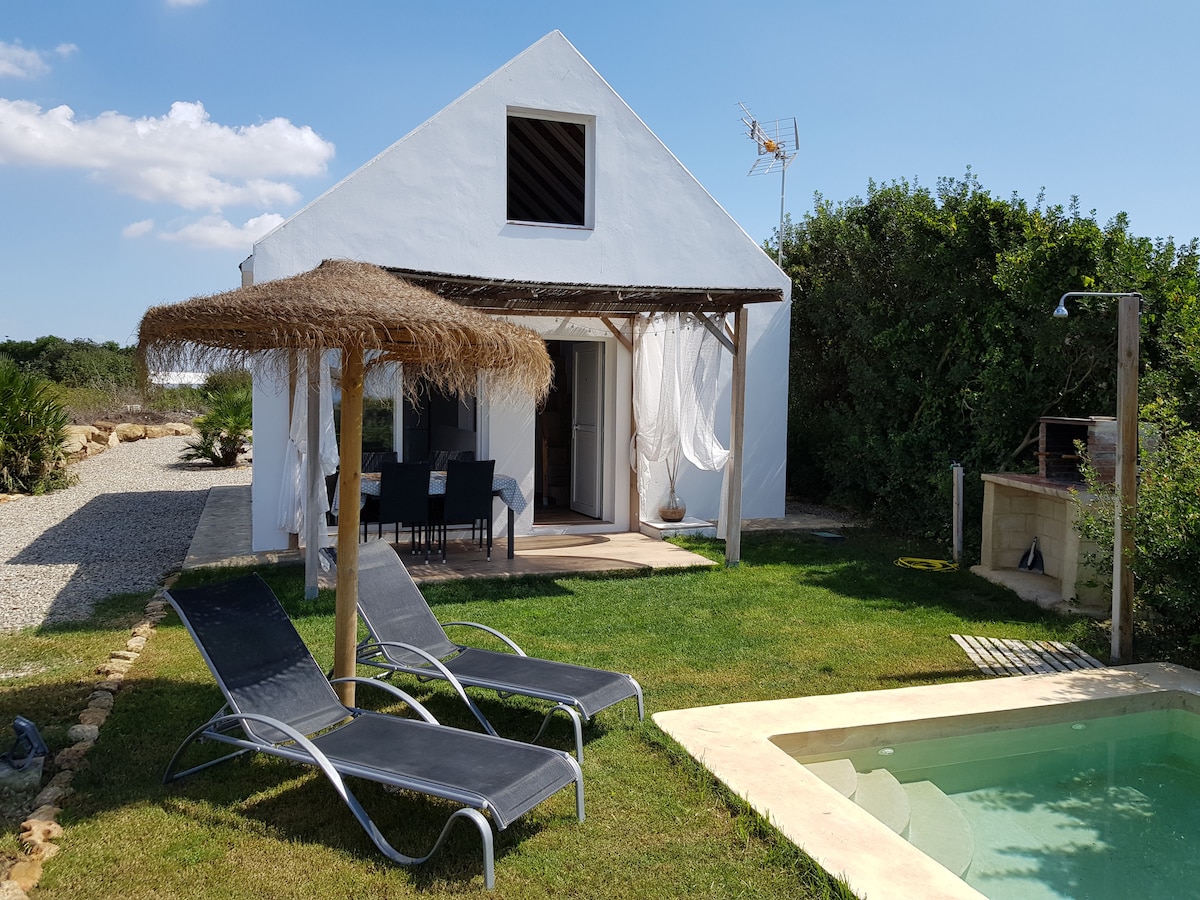 A white house with a thatched umbrella is situated in a green outdoor area. Two loungers are positioned on the lawn near a swimming pool. The entrance features glass doors that lead into the interior, enhancing the connection with the surrounding nature.