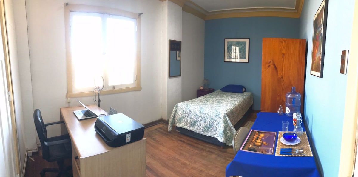 A simple bedroom features a light blue accent wall, a spacious bed adorned with a patterned blanket, and a desk with a printer. A large window allows natural light, while a wooden wardrobe and a side table with decorative items complete the room's furnishings.