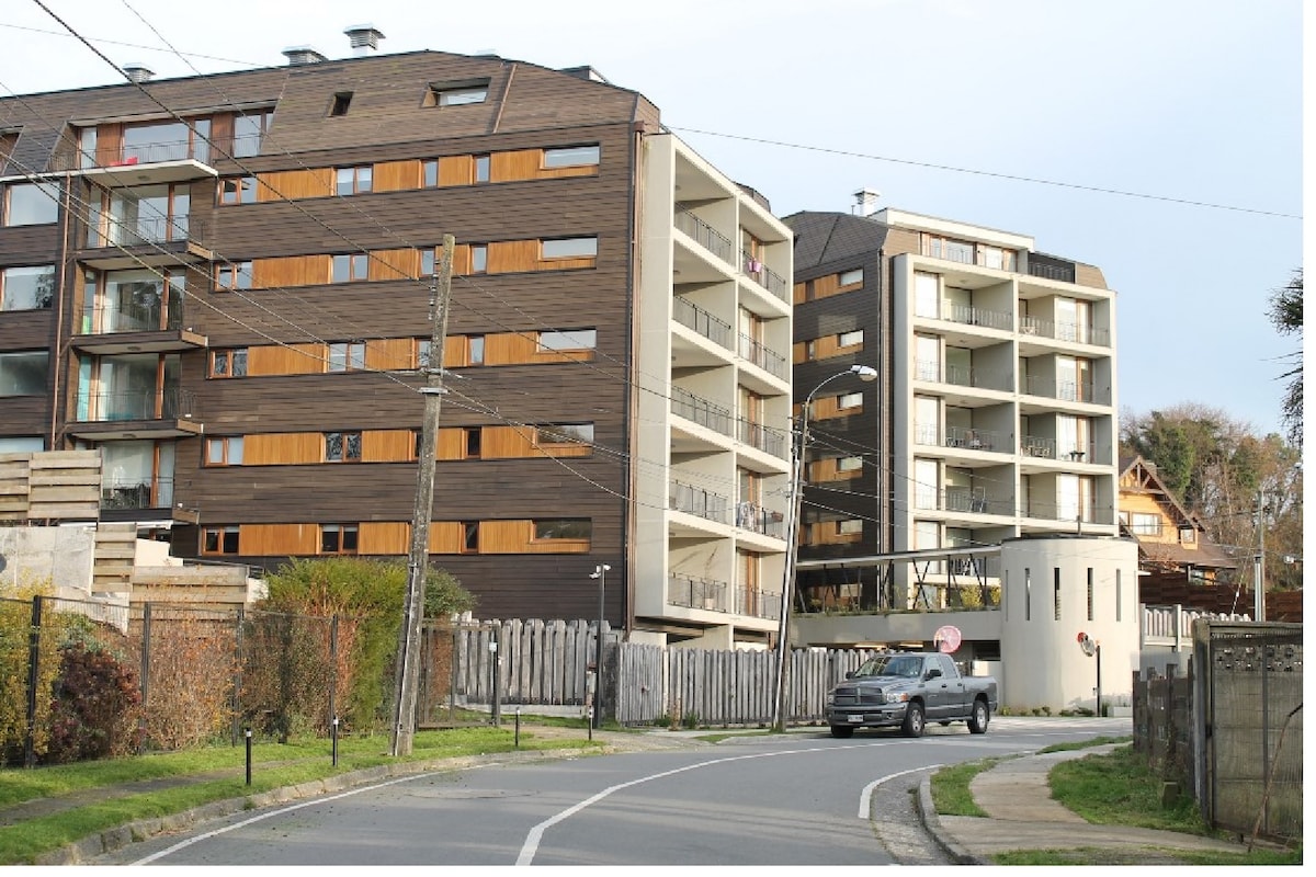 Two modern residential buildings with wooden facades are positioned along a road. Balconies with railings are visible on multiple levels. A circular structure can be seen in the foreground, with a gray vehicle passing by and a wooden fence in the background, adding to the community setting.