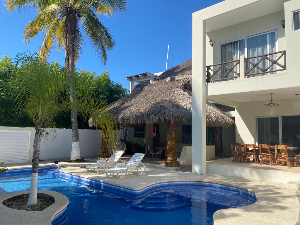 A swimming pool with a curved shape is complemented by two lounge chairs. A thatched-roof gazebo provides shade, surrounded by tropical trees. Nearby, a dining area features wooden furniture under a covered patio, with a modern two-story structure visible in the background.