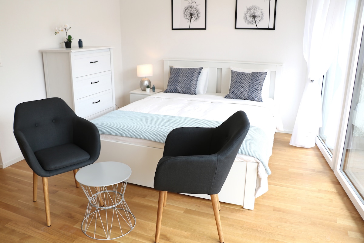 A cozy sleeping area features a spacious bed dressed in crisp white linens. Two modern armchairs flank a small table, creating a comfortable seating arrangement. Light-colored wood flooring and large windows enhance the bright and airy feel of the space.