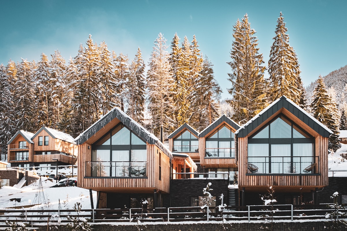 A modern chalet complex is surrounded by snow-covered trees. The wooden exterior features large glass windows that offer views of the winter landscape. The buildings are arranged at different heights, further enhancing the natural setting.