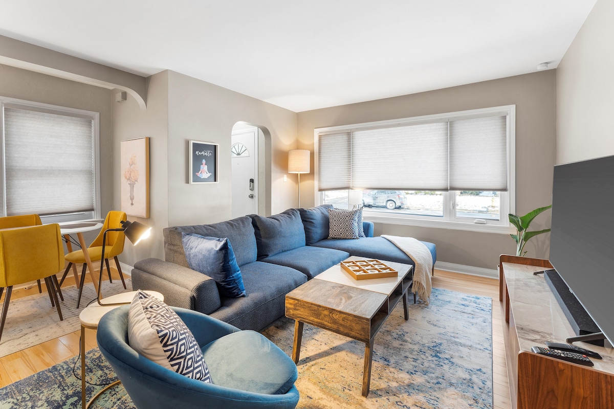 A living area features a large sectional sofa adorned with decorative pillows, complemented by a wooden coffee table. A stylish armchair and dining table for four are visible. Bright light filters through the blinds covering the windows, highlighting the warm color palette and wooden flooring.