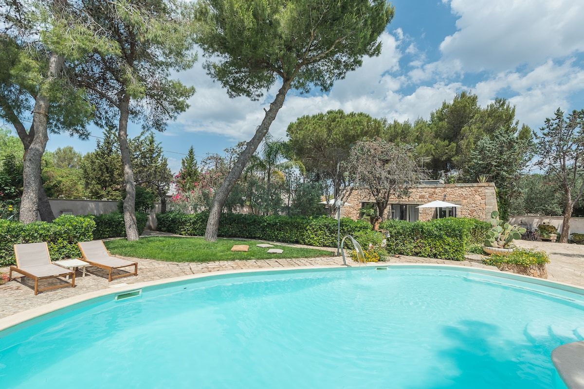 A serene pool area is surrounded by lush greenery, featuring two lounge chairs positioned beside the clear blue water. Tall trees provide shade, and a charming stone building can be seen in the background, blending harmoniously with the natural landscape.