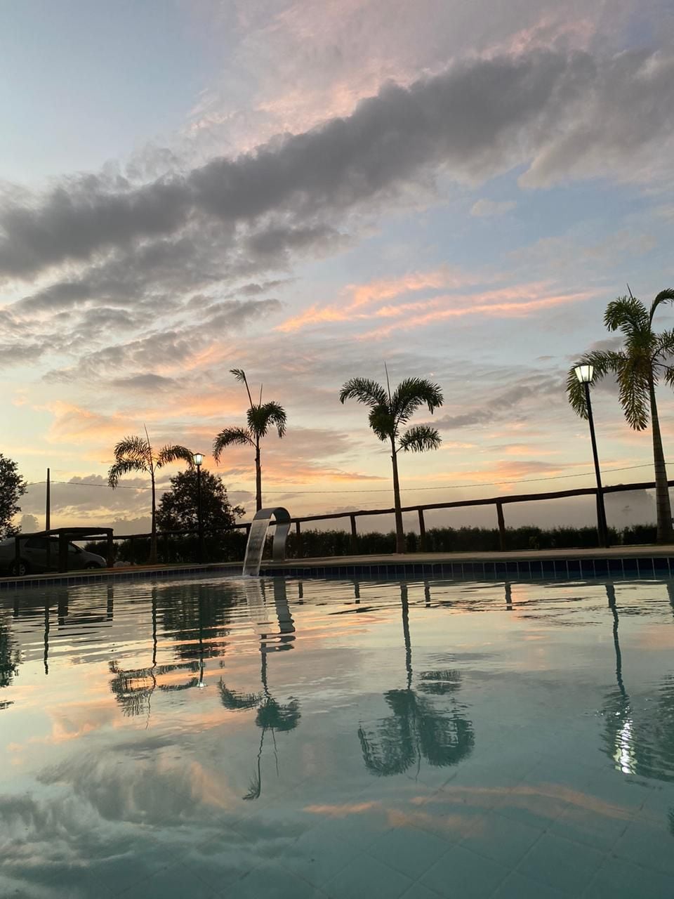 The swimming pool reflects the colorful evening sky, showcasing hues of orange and pink. Palm trees are visible along the poolside, and ambient lighting is provided by nearby lamps, creating a tranquil atmosphere for relaxation.