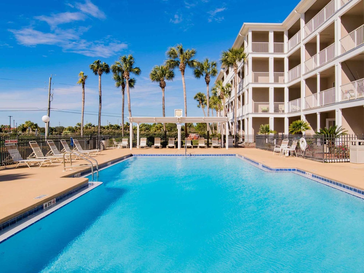 A spacious outdoor pool is surrounded by a sun deck, where lounge chairs are arranged under the clear blue sky. Palm trees are visible in the background, complementing the inviting atmosphere of the private gated community.