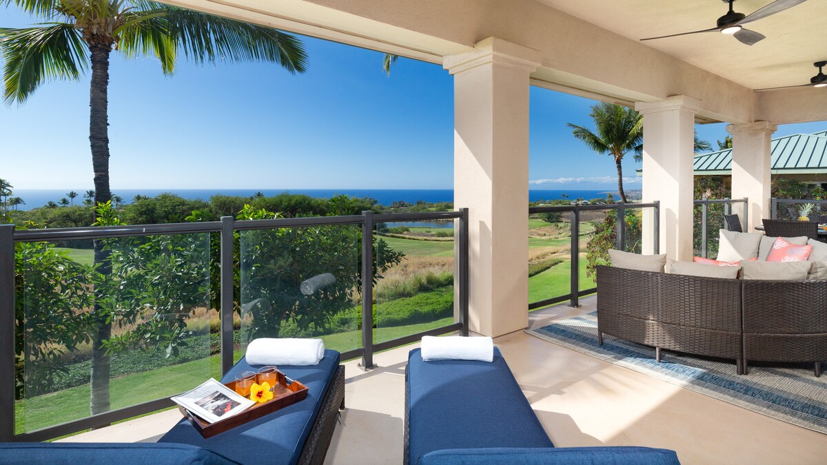 A spacious covered lanai provides comfortable lounge chairs overlooking the lush greenery of the golf course and the ocean in the distance. The view is enhanced by clear blue skies and palm trees creating a serene outdoor setting.