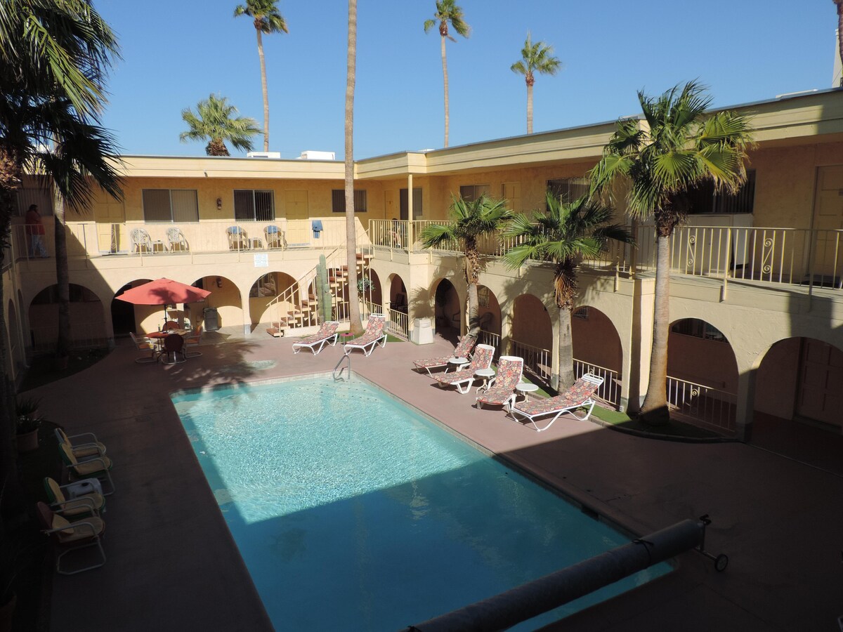 A large pool is surrounded by palm trees, with lounge chairs positioned around the shallow end. Arched walkways and a sunny patio area provide access to nearby rooms, creating a relaxed outdoor environment. The clear blue sky and warm sunlight enhance the inviting scene.