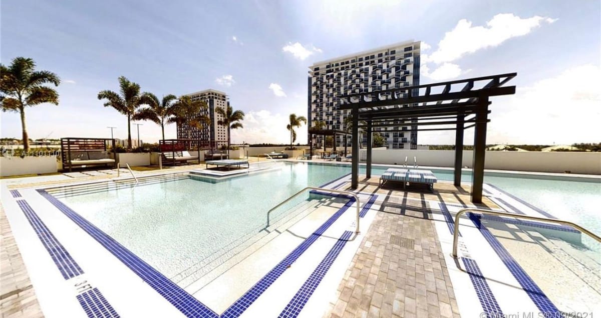 A spacious pool area is surrounded by palm trees, with lounge seating available under shaded structures. The water features a light turquoise color, emphasizing the modern design of the pool deck. The backdrop includes contemporary residential buildings, enhancing the vibrant outdoor setting.