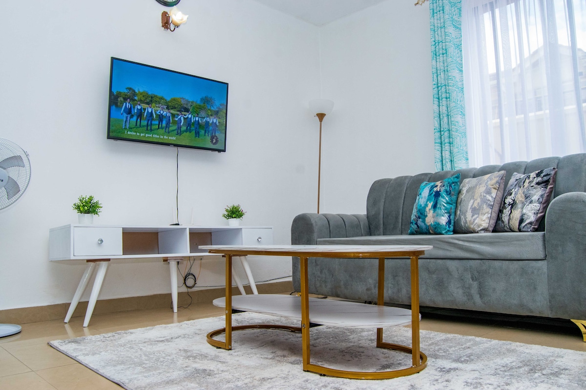 A living area features a grey sofa adorned with decorative cushions, complemented by a coffee table with a circular design. A wall-mounted flat-screen TV is visible above a white side table, while natural light filters through sheer curtains, illuminating a neutral-toned carpet.