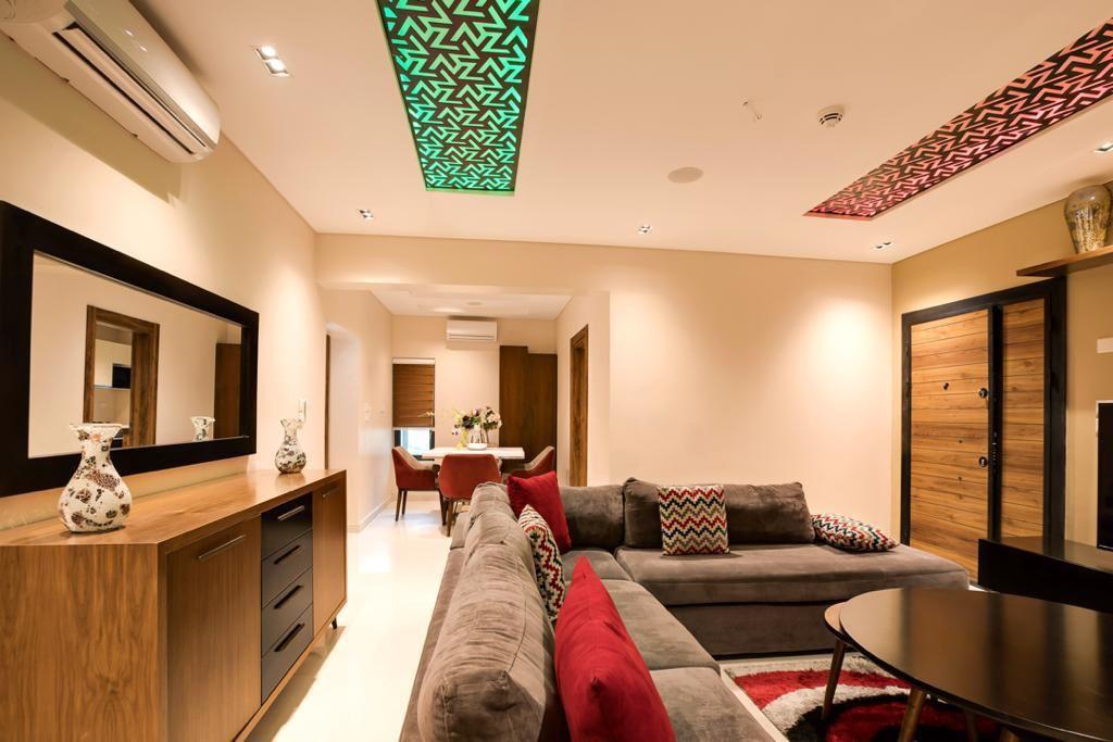 A modern living area is presented with stylish decor, featuring a spacious gray sectional sofa adorned with red and patterned cushions. Decorative wall lights in green and red illuminate the ceiling. A wooden sideboard and a round coffee table complement the space.