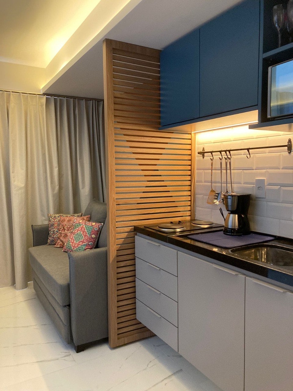 A compact kitchen area is shown, featuring a built-in counter with a sink and a coffee maker. Light-colored cabinets contrast with a blue upper cabinet. A cozy sofa is positioned nearby against a textured wooden partition, with decorative cushions adding a touch of color.