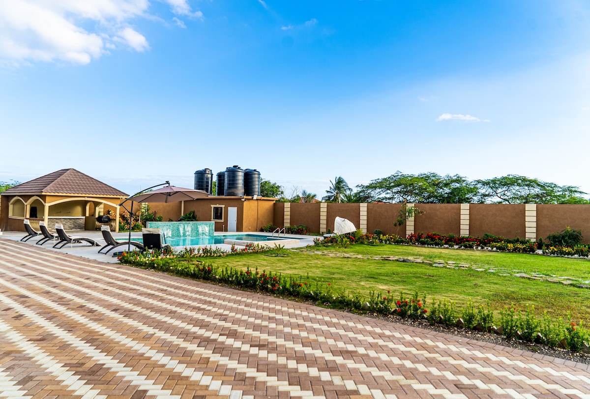 A well-maintained outdoor area is presented, featuring a crystal blue pool surrounded by a sun deck with several lounge chairs. A manicured lawn with colorful flower beds frames the space, while a shaded gazebo and water tanks are visible in the background.