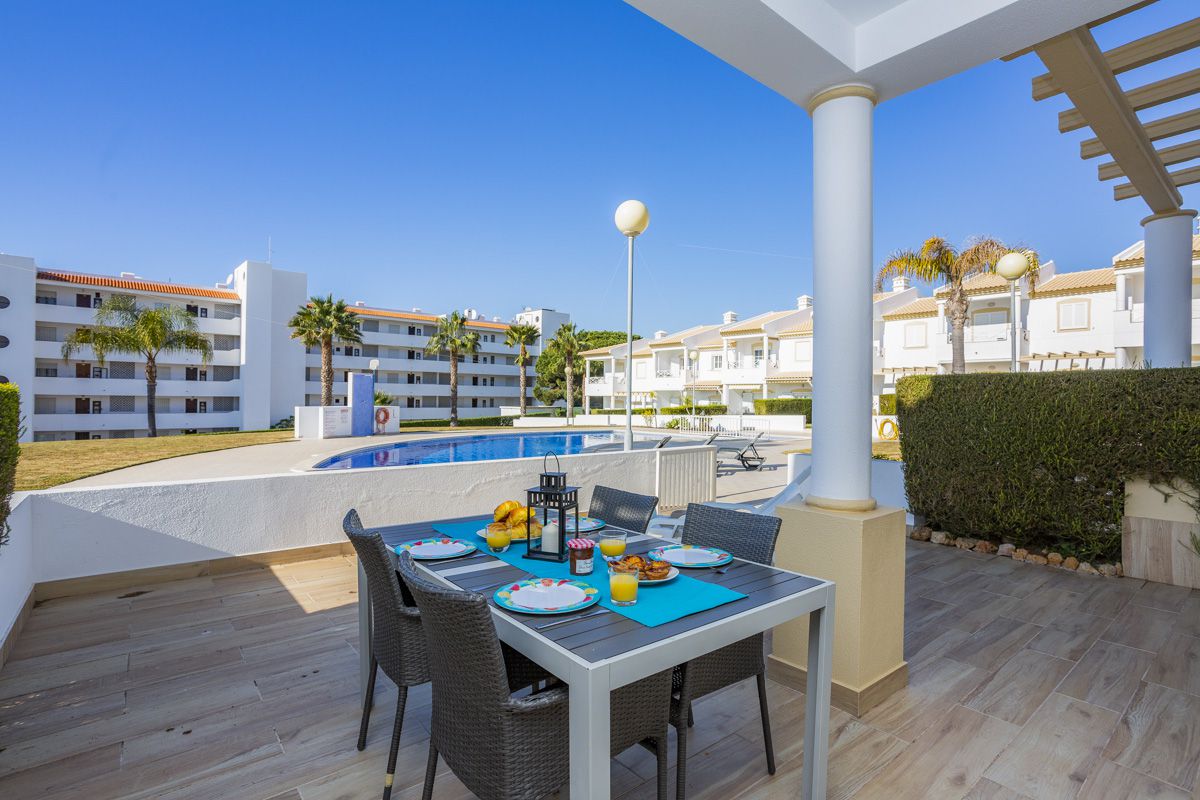 A private terrace features a dining table set for Al Fresco dining, with plates and fruit displayed. The view encompasses the communal swimming pool and garden area, highlighted by palm trees under a clear blue sky.