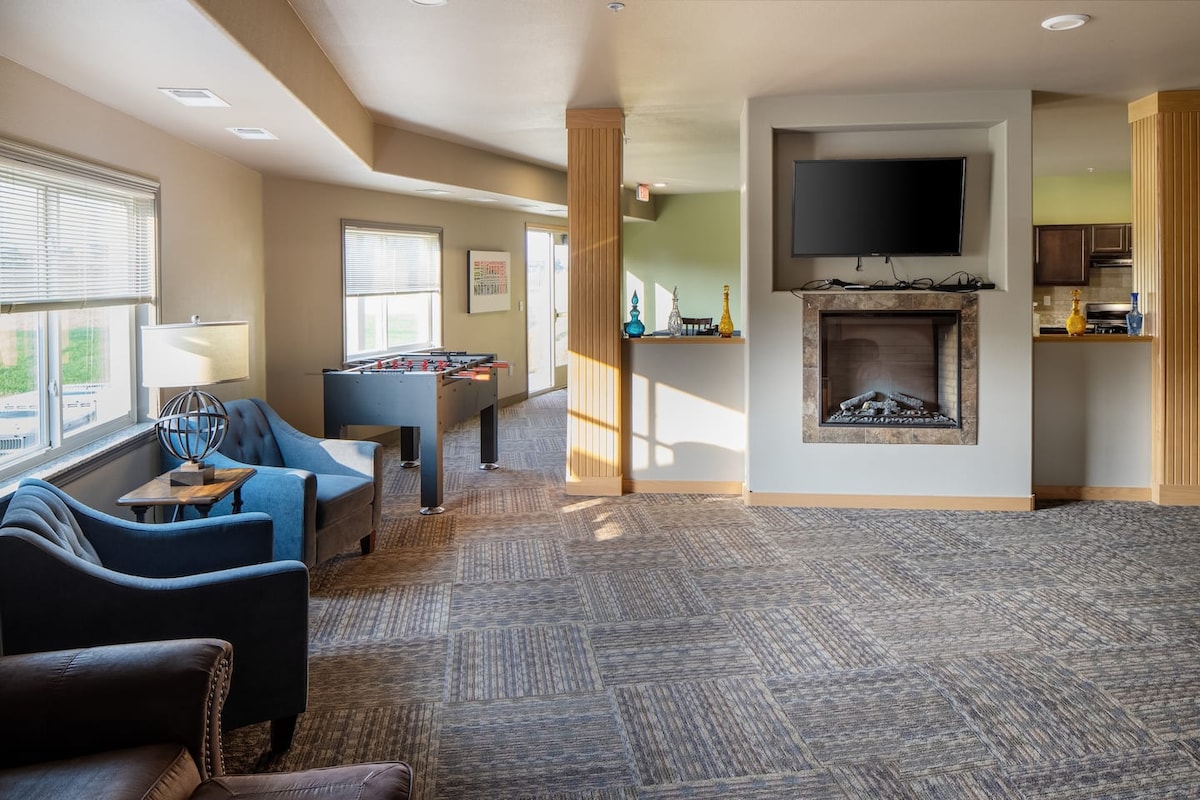 A common area is shown, featuring a cozy seating arrangement with blue and brown armchairs. A stylish fireplace is set beneath a wall-mounted TV. A foosball table is visible in the background, alongside a kitchenette area with dark cabinetry.