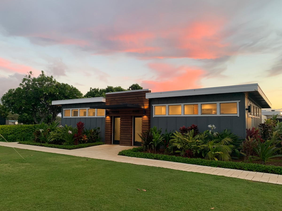 A modern condominium exterior is framed by lush tropical landscaping. The building features large windows and a welcoming entrance, with a path leading toward the front door. Soft colors from a sunset create a peaceful ambiance in the sky overhead.