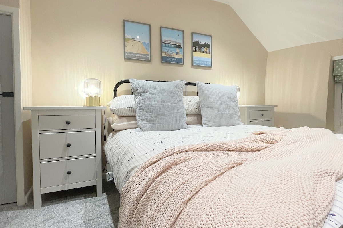 A cozy bedroom displays a comfortable double bed, adorned with light-colored pillows and a soft throw blanket. Two nightstands with drawers are positioned on either side, complementing the muted walls. Three artwork pieces depicting beach scenes are mounted above the bed, enhancing the serene ambiance.