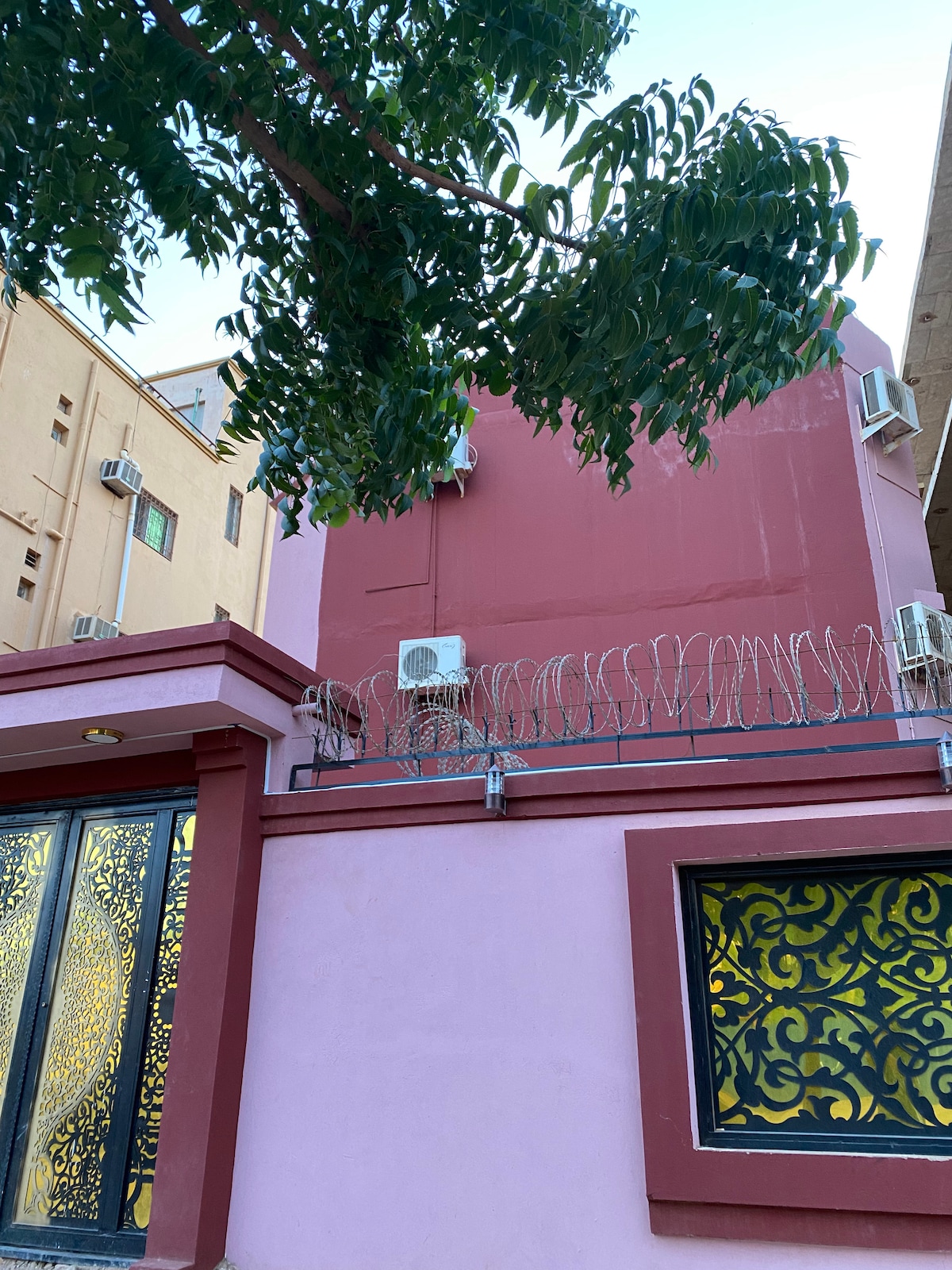 The exterior façade of a multi-story building is presented, showcasing a pink color scheme accented by decorative window frames in a contrasting dark tone. Security features, including barbed wire, are visible on the rooftop, and multiple air conditioning units are mounted on the walls.
