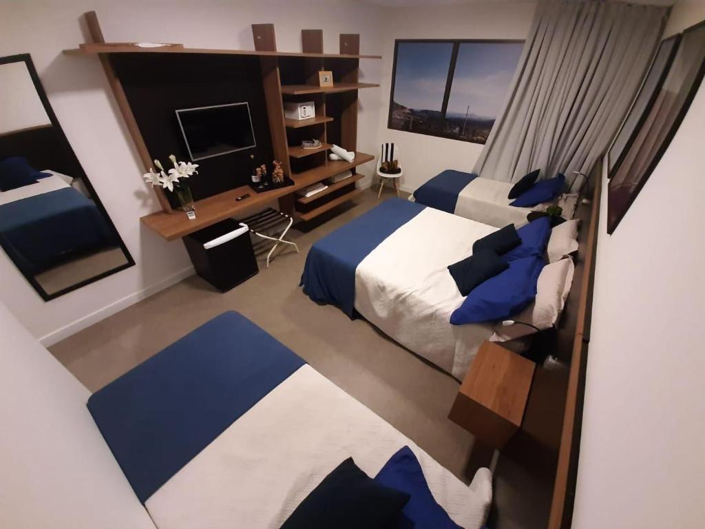 A well-organized bedroom features two spacious beds dressed in white and blue linens, complemented by a sleek wooden headboard. A modern television is mounted on the wall above a wooden console, which includes decorative items and additional cushions. Large windows allow natural light to illuminate the room.