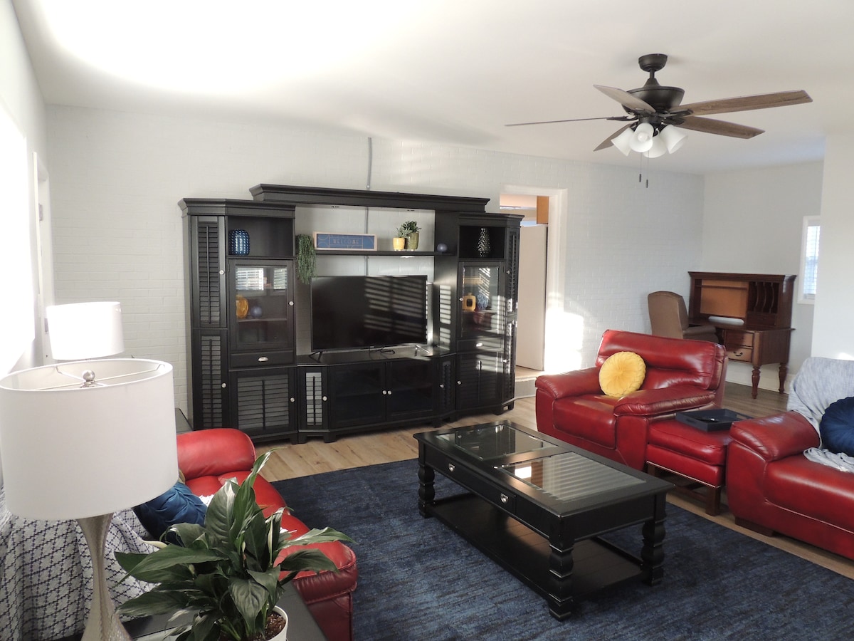 A spacious living area is furnished with red reclining sofas and a dark wood coffee table. A large entertainment unit housing a flat-screen television is centered against the wall. A ceiling fan and a floor lamp provide light, while a potted plant adds a touch of greenery.