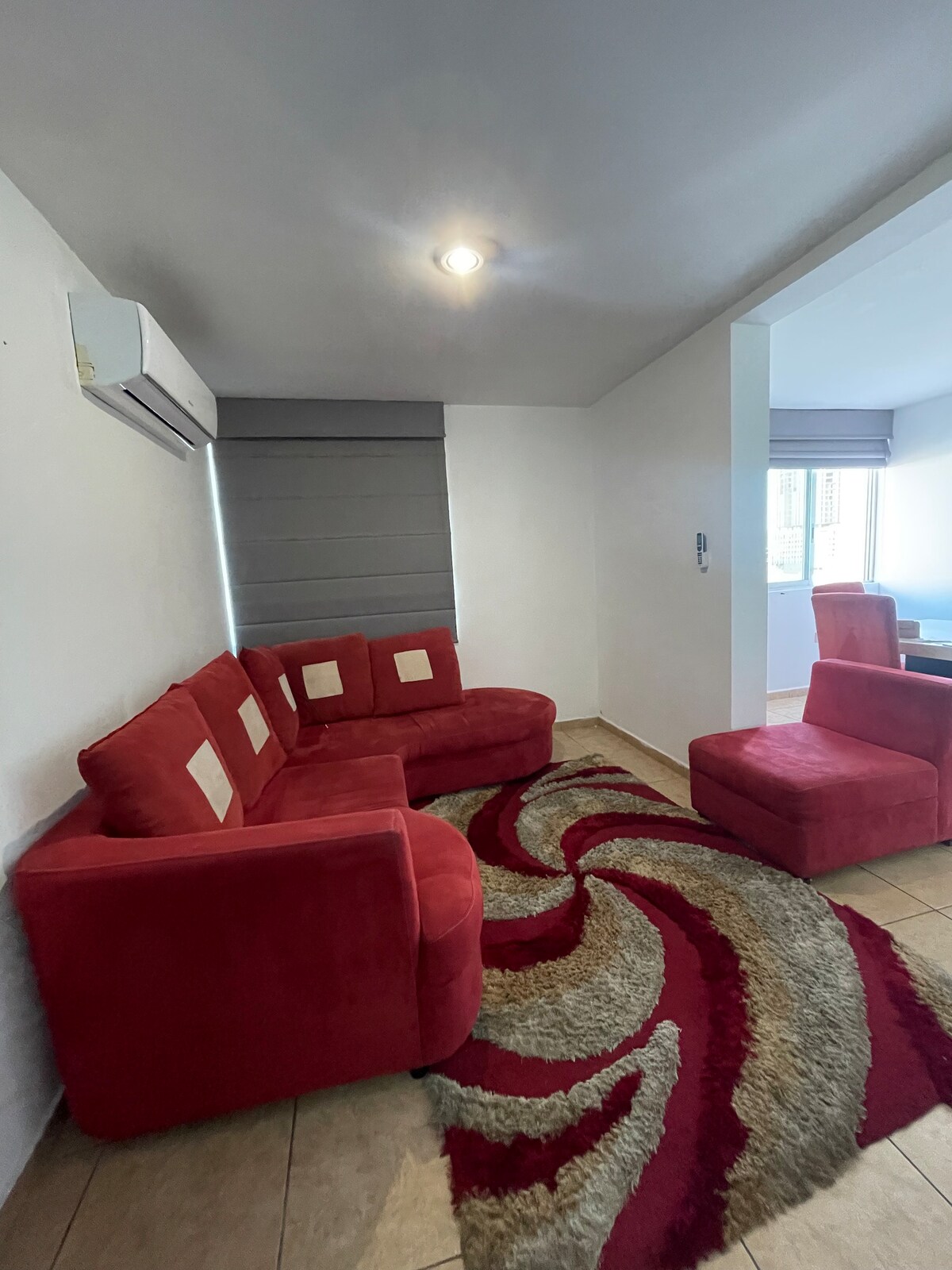 A comfortable seating area is furnished with a red sectional sofa and a matching chair. A patterned area rug adds texture to the space. Natural light enters through partially covered windows, creating a bright and inviting atmosphere.
