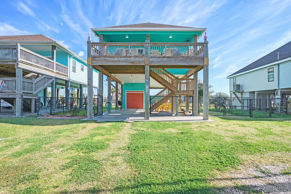 A two-story house with a vibrant green exterior is surrounded by a grassy area. The structure features a spacious wooden deck with stairs leading down. The lower level has an orange garage door, while the surroundings include glimpses of nearby homes and outdoor play areas.