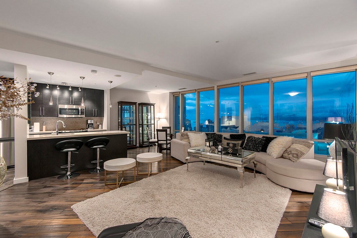 A spacious living area features large windows that offer expansive views of the city skyline. Elegant furniture is arranged on a soft area rug, while a comfortable sectional couch invites relaxation. The kitchen area is equipped with modern appliances, providing a stylish and functional space.