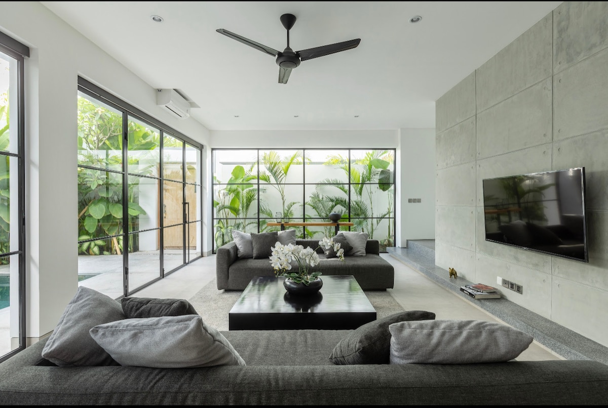 A modern living room features large glass windows, inviting natural light and views of greenery. A black coffee table sits at the center, surrounded by spacious dark sofas adorned with gray cushions. A flat-screen TV is mounted on the wall, enhancing the contemporary design.
