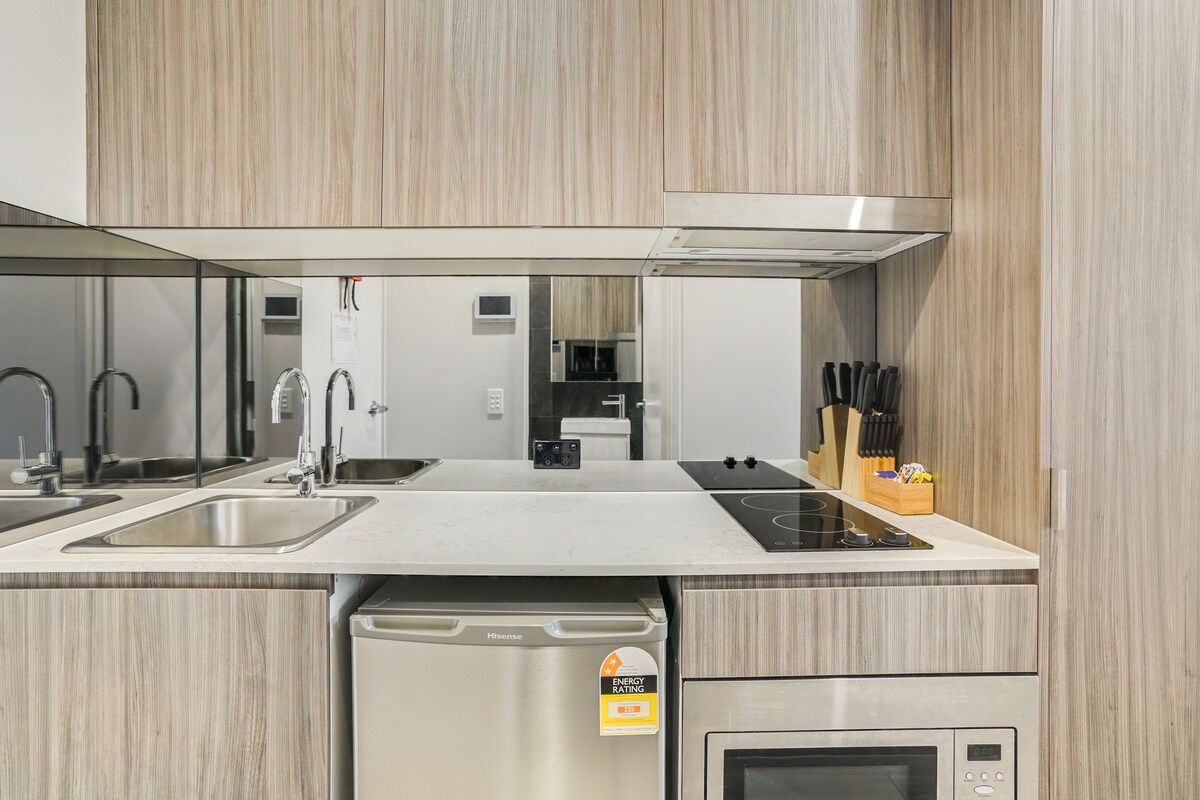 A modern kitchen is featured with sleek wooden cabinetry and a mirrored backsplash. Stainless steel appliances, including a refrigerator and microwave, are positioned alongside a two-basin sink and an electric cooktop, providing a functional cooking space.