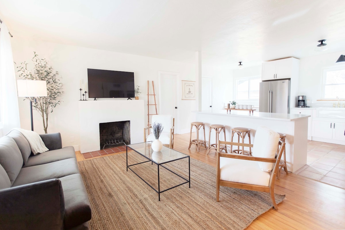A bright living area features a modern black couch and a cream accent chair. A sleek metal coffee table sits on a textured rug, while a wall-mounted TV is positioned above a minimalist fireplace. Natural light flows through large windows, enhancing the spacious feel.