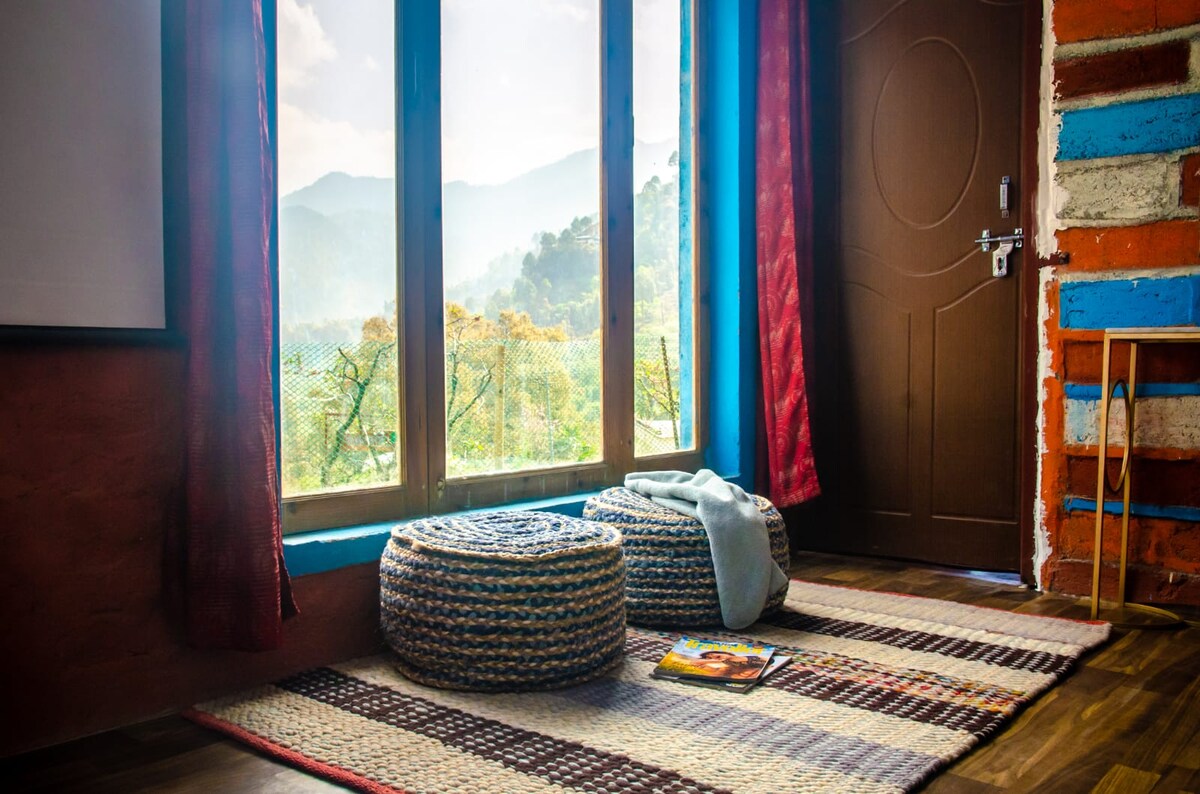 A tranquil sitting area is created with woven poufs arranged on a textured rug. Large windows allow natural light to fill the space, revealing scenic views of rolling hills in the distance, framed by vibrant curtain drapes.