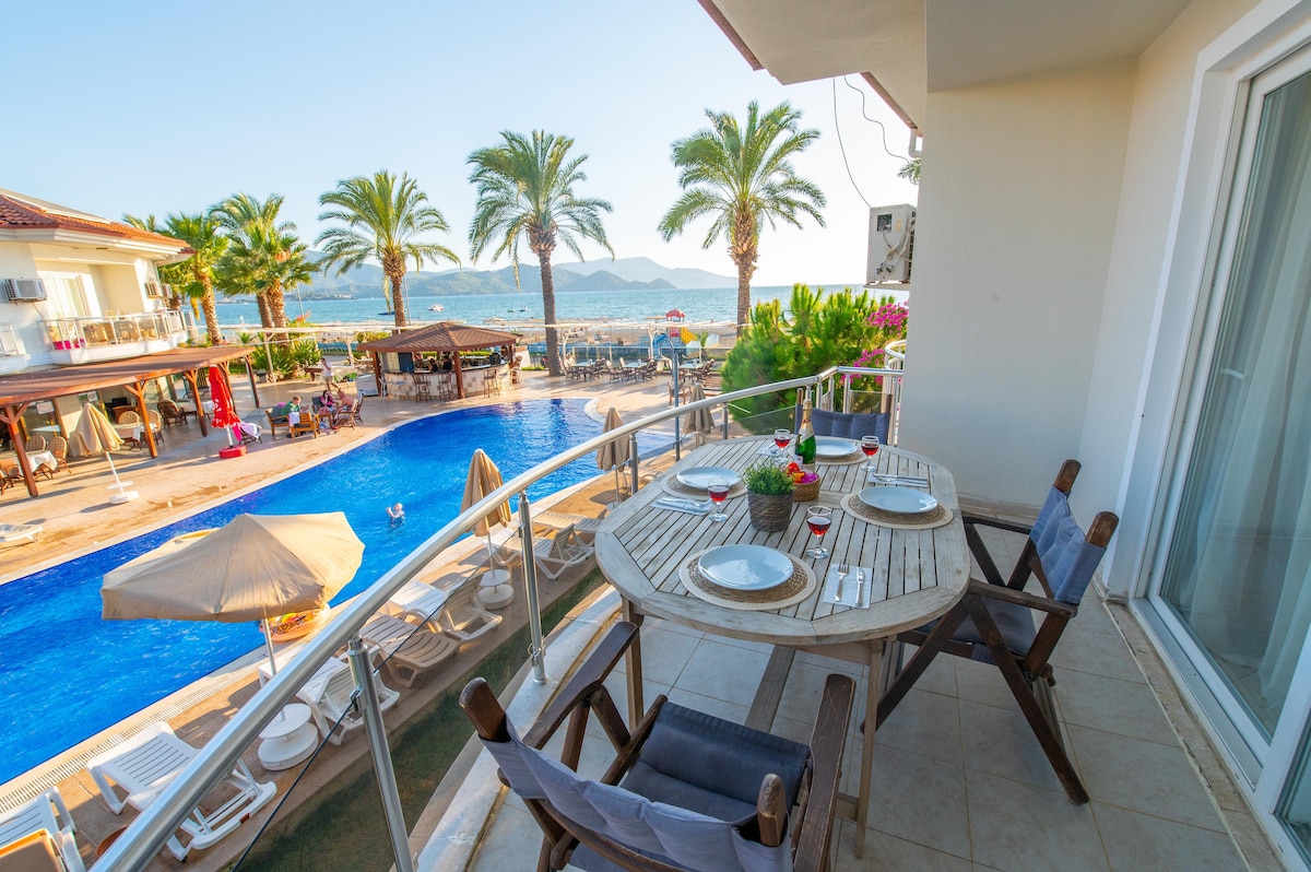 A balcony table is set for dining, featuring dishes and glasses overlooking a pool surrounded by palm trees. Sun loungers and umbrellas are positioned near the water, with distant mountains visible against the clear sky.