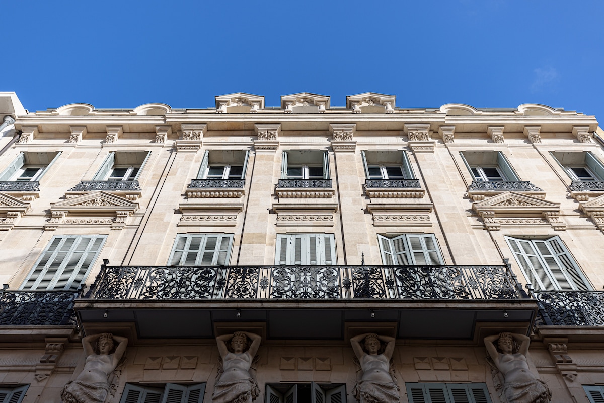 The exterior of a historic building is highlighted, showcasing intricate architectural details such as decorative moldings, wrought-iron balconies, and tall windows. The façade features classical sculptures supporting the balcony, set against a clear blue sky.