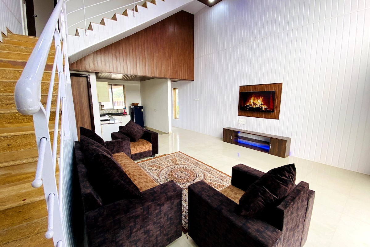 A spacious living area showcases two comfortable sofas facing a modern flat-screen TV mounted on the wall. The room features a plush area rug on tile flooring, with natural light streaming in from large windows above. A staircase with a wooden railing adds architectural interest.