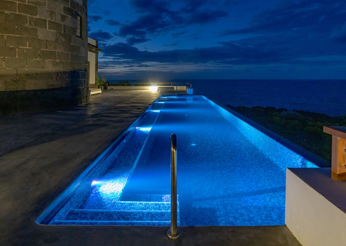 An illuminated infinity pool reflects the deep blue of the ocean under a twilight sky, with soft lighting enhancing the serene atmosphere. The pool's edge appears to blend seamlessly with the horizon, offering a tranquil space for relaxation.