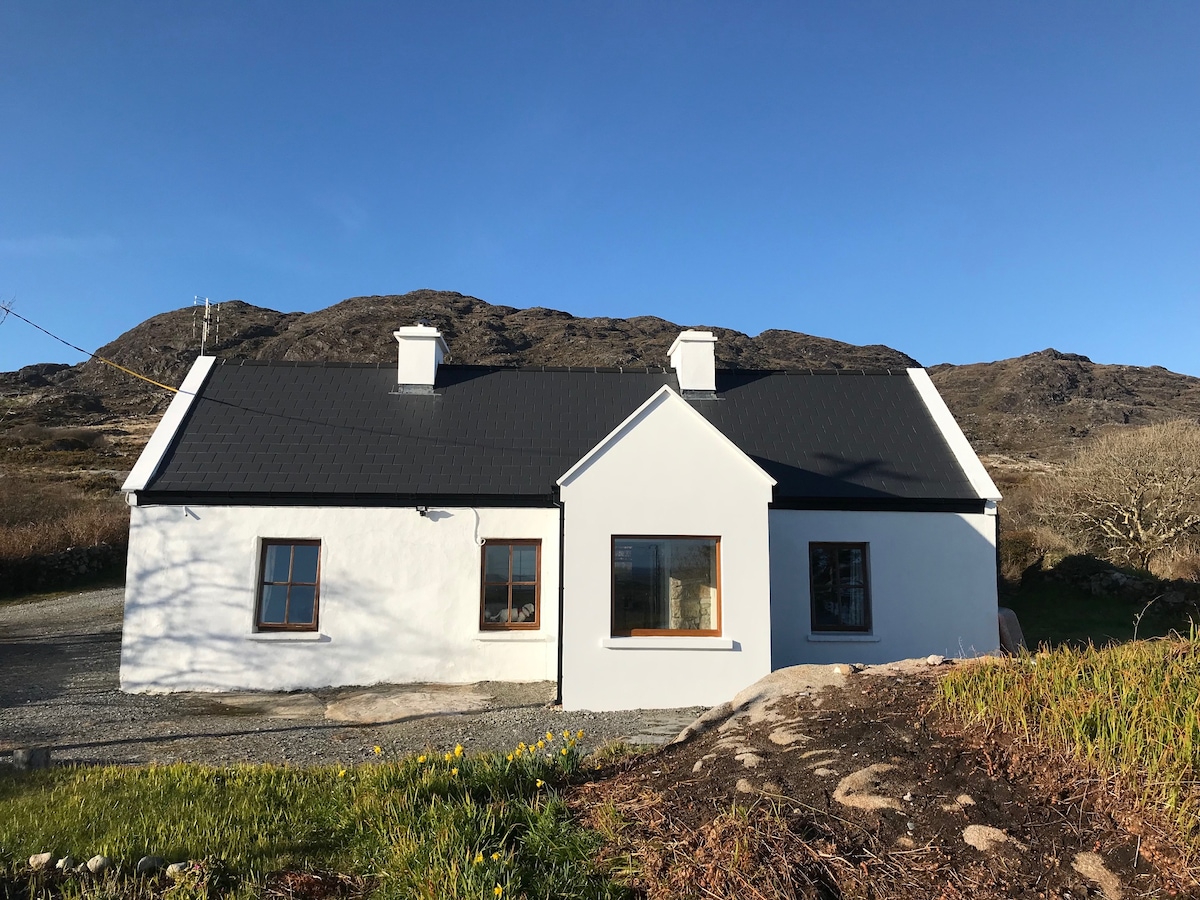 The front view of the cottage displays a traditional design with a white exterior and a dark, sloped roof. A gravel pathway leads to the entrance. The surrounding landscape features rugged terrain and distant mountains under a clear blue sky.