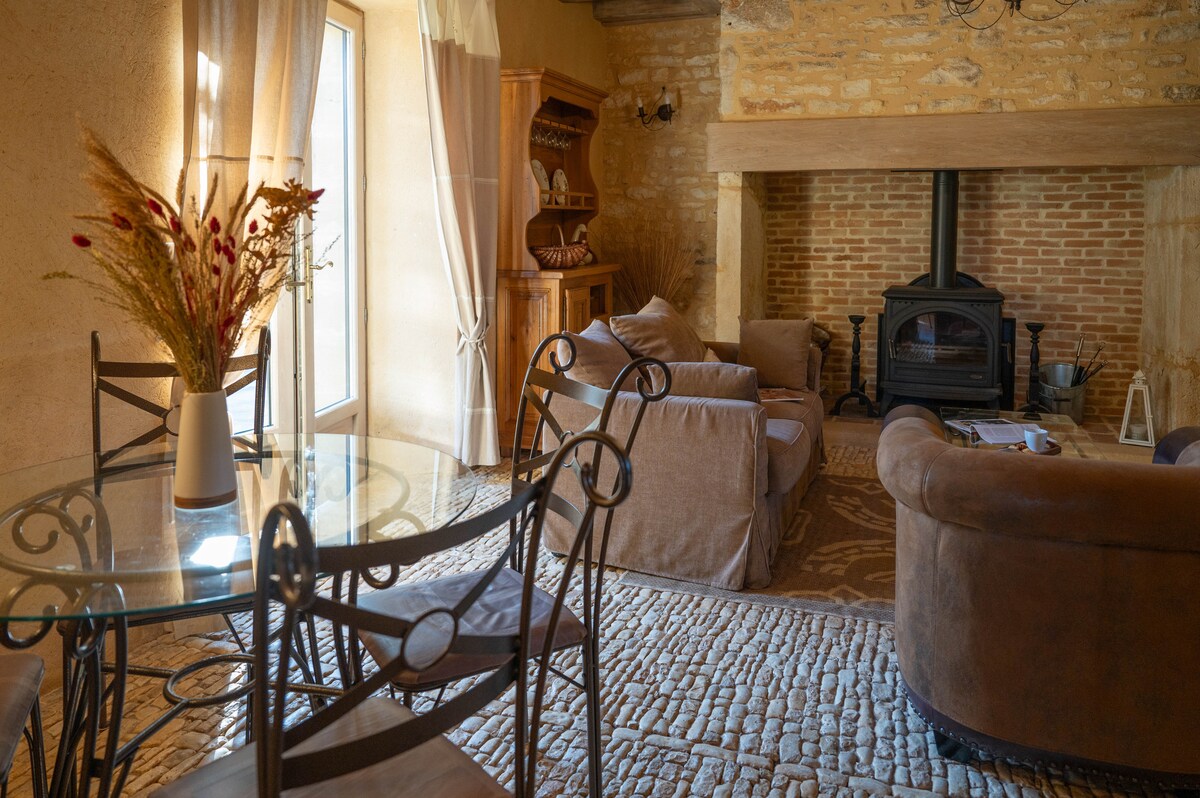 A cozy living area is depicted with a round dining table featuring black metal chairs and a decorative arrangement of dried flowers. A comfortable seating arrangement surrounds a wood-burning stove, with natural light filtering through large windows, illuminating the warm stone walls.